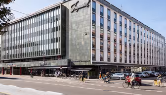 En moderne bygning i flere etasjer med et stort skilt som viser Imperial. Fasaden inkluderer mange vinduer. Det er en restaurant på gateplan med uteservering, og folk går og sykler i nærheten på en solrik dag.