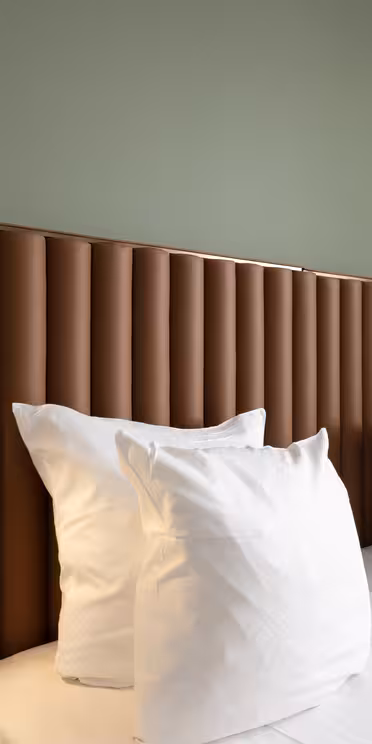 Moderne hotellrom med to pent oppredde enkeltsenger med hvitt sengetøy og flere puter. Sengegavlen er en polstret brun design med integrerte svarte leselys. Veggene er malt i en myk grønn, og beige gardiner er delvis synlige.