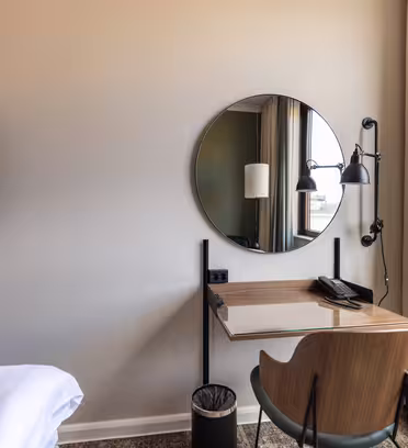 Et minimalistisk hotellrom med skrivebord i tre med rundt speil, moderne lampe og telefon. En elegant svart TV er montert på veggen. En komfortabel stol og søppelbøtte fullfører oppsettet. Lyset strømmer inn gjennom et stort vindu med beige gardiner.