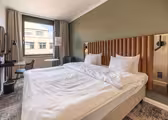 Hotellrom med en stor seng med hodegjerde i tuftet skinn. Det er et vindu med gardiner, en grønn aksentvegg, et lite skrivebord med en stol, et sidebord med en lampe og moderne innredning. Et mykt teppe dekker en del av gulvet.