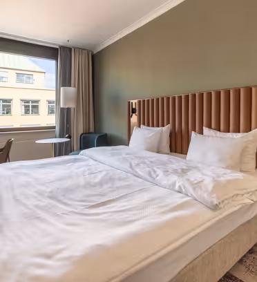 Hotellrom med en stor seng med hodegjerde i tuftet skinn. Det er et vindu med gardiner, en grønn aksentvegg, et lite skrivebord med en stol, et sidebord med en lampe og moderne innredning. Et mykt teppe dekker en del av gulvet.