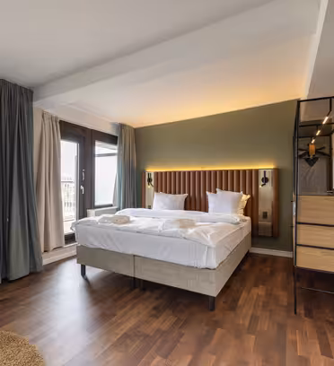 Et moderne hotellrom med en pent oppredd dobbeltseng med hvitt sengetøy. Rommet har tregulv, TV på veggen, høy lampe og store vinduer med gardiner. Det er et lite rundt bord og en stilig bokhylle ved siden av sengen.