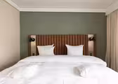 Et godt opplyst hotellrom har en pent laget king-size-seng med hvitt sengetøy og to puter. Sengegavlen er polstret og veggen er malt grønn. To veggmonterte lys flankerer sengen, og en gardin dekker delvis et vindu til høyre.