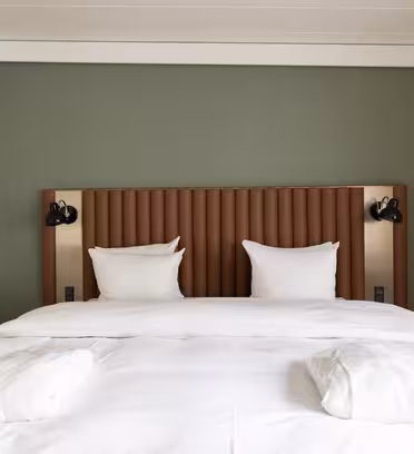 Et godt opplyst hotellrom har en pent laget king-size-seng med hvitt sengetøy og to puter. Sengegavlen er polstret og veggen er malt grønn. To veggmonterte lys flankerer sengen, og en gardin dekker delvis et vindu til høyre.