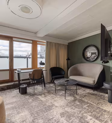 Et moderne hotellrom med stort vindu og utsikt over byen. Den har et rundt speil, en lysegrå sofa, en svart stol, et lite rundt bord og en TV montert på veggen. Rommet har nøytral-tonet innredning og et teksturert teppe.