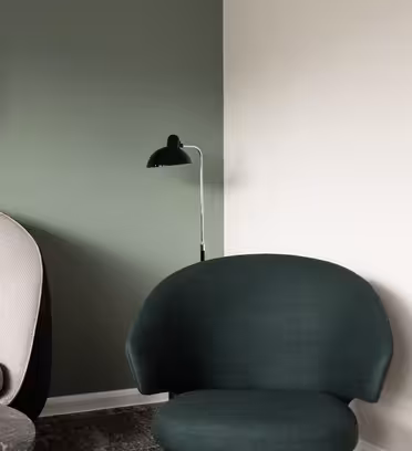 Et moderne interiør har en grønn lenestol, en beige sofa og et rundt speil på veggen. En svart gulvlampe står i nærheten. Veggene er malt i kontrasterende nyanser av grønt og hvitt.