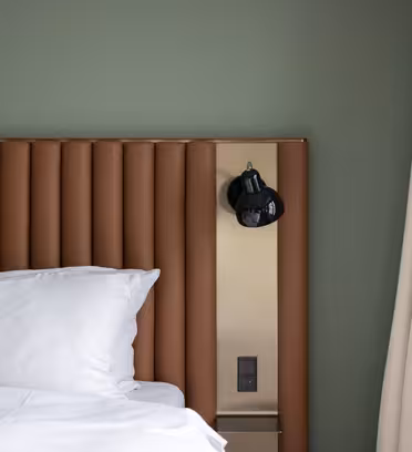 Et moderne hotellrom med en seng med hvite puter og laken. Sengegavlen er brun og vertikalt rillet. En sort vegglampe er montert på sengegavlen, ved siden av en beige vegg og beige gardiner. Veggen er malt grønn.