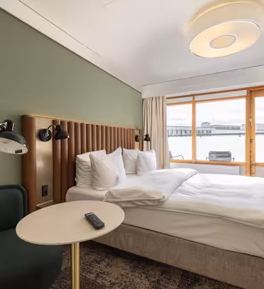 Et hotellrom med en pent oppredd dobbeltseng. Rommet har en grønn aksentvegg og et rundt bord med fjernkontroll for fjernsynet. En plysjstol sitter i nærheten. Store vinduer gir naturlig lys og utsikt over bybildet.