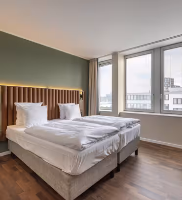 Moderne hotellrom med to pent oppredde enkeltsenger mot en grønn aksentvegg. Store vinduer til høyre gir utsikt over byen. Rommet har tregulv, en koselig sittegruppe med en blågrønn stol og bord, og dempet belysning for atmosfære.