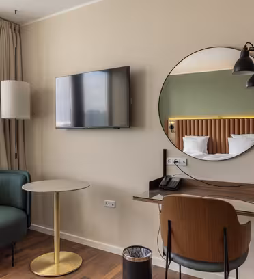 Moderne hotellrom med et lite skrivebord, stol, rundt speil og veggmontert TV. En koselig lenestol og rundt bord er nær vinduet. Sengen er delvis synlig med grønn- og tredekor. En lampe henger ved siden av skrivebordet.