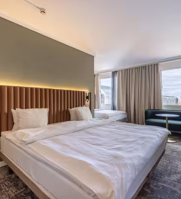 Et moderne hotellrom med en stor seng, beige og grønn innredning og et teksturert teppe. Et sittehjørne med grønn sofa og rundt bord er ved vinduet, som gir utsikt over bybildet. Et skrivebord og en gulvlampe er til høyre.