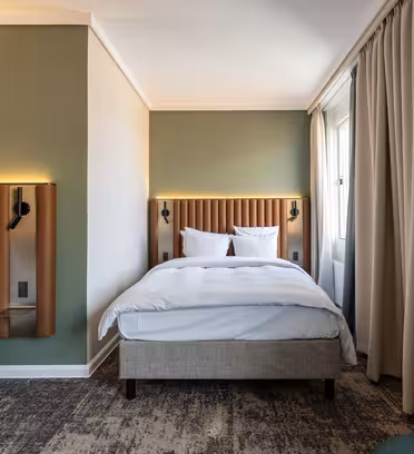 Hotellrom med to senger, hver med polstret sengegavl og skarpt hvitt sengetøy. Rommet har en grønn aksentvegg, beige gardiner og mønstret teppe. En koselig grønn stol er synlig i nedre høyre hjørne, og rommet er godt opplyst.