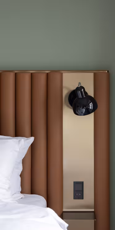 Soveromsinteriør med en seng med hvite puter og laken. Sengegavlen er trukket i brunt, og det er en innebygd lampe på et panel ved siden av. En gardin henger til høyre mot en dempet grønn vegg.