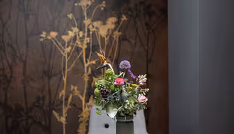 En vase med en mangfoldig blomsterbukett, inkludert en paradisfugl og lilla allium, står på et bord. Bakgrunnen har en mørk vegg med en silhuettdesign av høye planter.