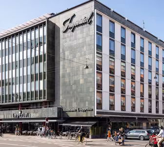 En moderne bygning i flere etasjer med et stort skilt som viser Imperial. Fasaden inkluderer mange vinduer. Det er en restaurant på gateplan med uteservering, og folk går og sykler i nærheten på en solrik dag.