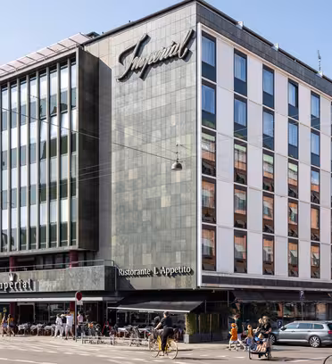 En moderne bygning i flere etasjer med et stort skilt som viser Imperial. Fasaden inkluderer mange vinduer. Det er en restaurant på gateplan med uteservering, og folk går og sykler i nærheten på en solrik dag.