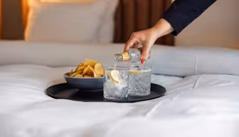 En person heller en klar drikk fra en liten flaske over i et glass med is og sitron på et brett, ved siden av en bolle med potetgull på en pent oppredd seng.