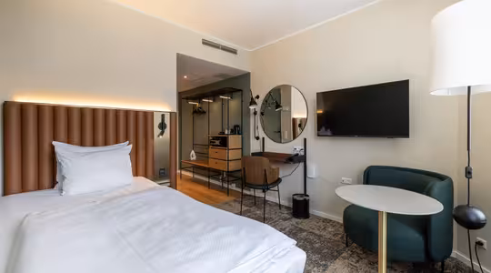 Moderne hotellrom med pent oppredd seng, polstret hodegavl, veggmontert TV, rundt speil, skrivebord med stol, grønn lenestol, rundt bord og dempet belysning. Garderobe og inngangsparti er synlige i bakgrunnen.