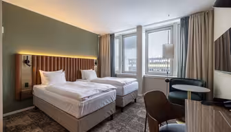 Moderne hotellrom med to enkeltsenger, polstret hodegavl, grønn aksentvegg, store vinduer med utsikt over byen, skrivebord, stol og et lite rundt bord med lenestol. Nøytrale toner og moderne innredning.