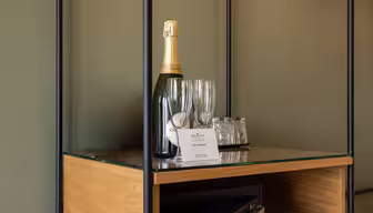 En flaske musserende vin, to tomme champagneglass og et brett med to glass er plassert på en glasshylle med trebunn. Et lite kort er plassert foran gjenstandene.
