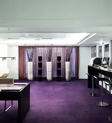 Moderne kontorinteriør med elegant design. Funksjonene inkluderer et skrivebord med beige stoler til venstre, en rad med datamaskiner med barkrakker til høyre, store vaser med grener i bakgrunnen og et lilla teppe overalt.