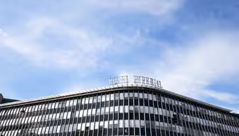 En moderne fleretasjes glass- og stålbygning med buet design mot en blå himmel. Vinduene reflekterer skyene, og det er en liten del av skilting på taket.