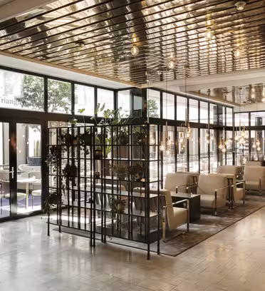 Romslig moderne kafé med glassvegger og skillevegger i metallramme. Inne er komfortable beige stoler og små bord på flislagt gulv. Taklamper reflekterer metallaksenter og skaper en varm atmosfære. Planter legger til grønt i hele plassen.
