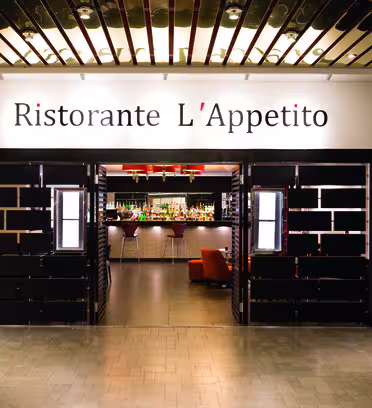 Inngang til "Ristorante L'Appetito" med svart og trepanel. Innvendig er en godt opplyst bar synlig med ulike flasker og glass. Interiøret har moderne innredning med varme toner og sitteplasser.