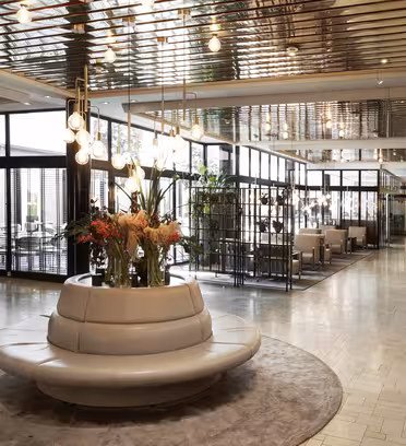 Moderne hotellobby med en sirkulær grå sittegruppe og et blomstret midtpunkt. Overhead henger unike lysarmaturer fra et speiltak. Konferanserom er synlige i bakgrunnen, og et høyt katalogskilt vises til høyre.