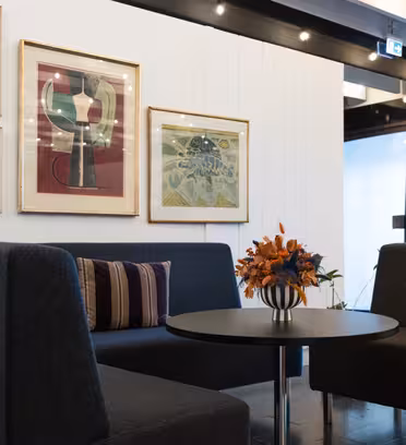 Et moderne salongområde har et rundt bord med en vase med oransje blomster, omgitt av mørkegrå stoler. De hvite veggene viser fargerik abstrakt kunst. Taksporlys er synlige, og skaper en koselig atmosfære.