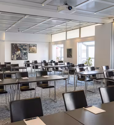 Et moderne konferanserom med rader med svarte stoler og bord pent ordnet. Hvert bord har en notisblokk og penn. Rommet er godt opplyst med store vinduer og abstrakt kunst på veggene. En projektor er montert i taket.