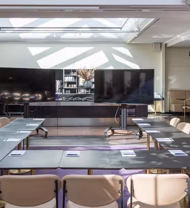 Et godt opplyst konferanserom med et arrangement av U-formede bord, beige stoler og to store skjermer foran. Sollyset strømmer gjennom et takvindu, og kaster mønstre på gulvet.