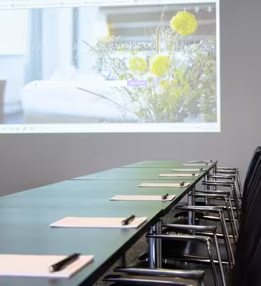 Et konferanserom med et langbord dekket med notatblokker og penner. I bakgrunnen viser en projektor et uskarpt bilde av en nettside med en bukett blomster. Rommet er moderne med minimalistisk design.