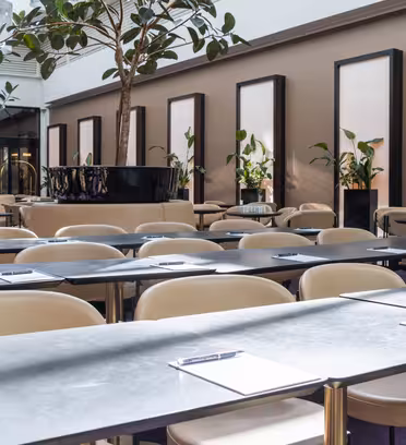 Konferanserom med flere rader med langbord og beige stoler, hvert bord har en utklippstavle og penn. Store potteplanter og høye speil pryder bakgrunnen. Rommet er godt opplyst med naturlig lys fra store vinduer.