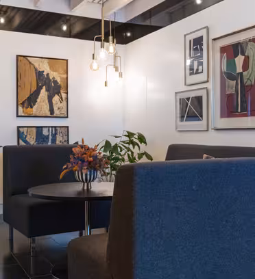 Moderne salong med blå sofaer, et rundt bord og et fargerikt blomsterarrangement. Veggene er utsmykket med abstrakt kunst. Hengende pendellamper gir en varm glød. Potteplanter forsterker den moderne innredningen.