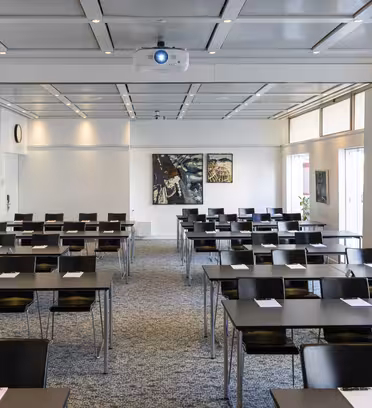 Et tomt konferanserom med rader med sorte stoler og bord arrangert i klasseromsstil. Hvert bord har en notisblokk og penn. En projektor er montert i taket, og det er kunstverk på veggene. Store vinduer langs høyre side.