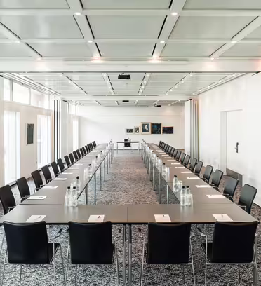 Et romslig konferanserom med to langbord arrangert i U-form. Hvert bord har svarte stoler og er dekket med notatblokker og flaskevann. Rommet er godt opplyst med store vinduer, hvite vegger og flere innrammede kunstverk i bakgrunnen.