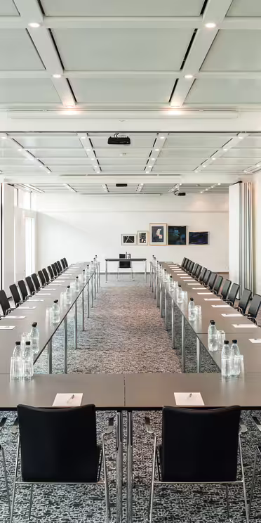 Et romslig konferanserom med to langbord arrangert i U-form. Hvert bord har svarte stoler og er dekket med notatblokker og flaskevann. Rommet er godt opplyst med store vinduer, hvite vegger og flere innrammede kunstverk i bakgrunnen.