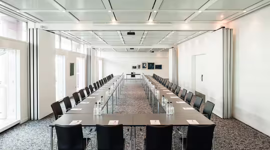 Et romslig konferanserom med to langbord arrangert i U-form. Hvert bord har svarte stoler og er dekket med notatblokker og flaskevann. Rommet er godt opplyst med store vinduer, hvite vegger og flere innrammede kunstverk i bakgrunnen.