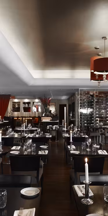 Moderne restaurantinteriør med bord i mørkt tre, sitteplasser i svart skinn og tente stearinlys. Røde lamper og fargerike kunstverk pryder rommet. En utstilling for vin i glass langs den ene veggen, og bidrar til den elegante stemningen.