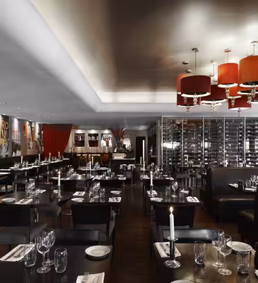 Elegant restaurantinteriør med dempet belysning, med moderne svarte bord dekket med glass og hvite servietter. Røde lysekroner henger i taket, og et stort vinstativ og fargerike veggmalerier pryder veggene.
