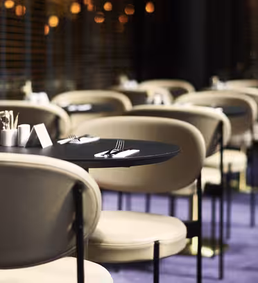 Moderne restaurantinteriør med rader med små runde bord dekket med bestikk på mørke flater. Beige polstrede stoler omgir hvert bord. Myk belysning og et lilla teppe skaper en varm og innbydende atmosfære.