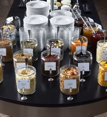 Et utvalg av frokostblandinger og toppingsglass med skjeer på en frokostbuffé. Bak glassene står stabler med tallerkener og boller, samt beholdere med forskjellige krydder og pålegg, satt på en mørk benke.