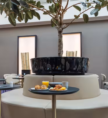 Et moderne kaféinteriør med et sentralt pottetre omgitt av sirkulære sitteplasser. Små bord er dekket med kopper og tallerkener med mat, inkludert bakverk og et glass appelsinjuice. Minimalistisk veggdekor med bambusaksenter komplementerer innstillingen.