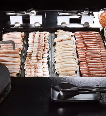 Et bufféoppsett med et utvalg av kjøtt ordnet pent i rader, inkludert baconstrimler og pølser, sammen med skåler med skivede agurker, tomater og rød paprika. Sorte serveringsredskaper og tallerkener er plassert i forgrunnen.
