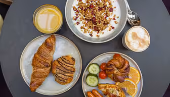 En frokost på et rundt bord med en bolle med yoghurt med granola og bær, et glass appelsinjuice, en cappuccino, en tallerken med en croissant og bakverk, og en annen tallerken med frukt, grønnsaker, ost og pølser.