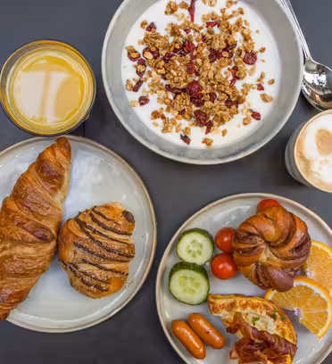 En frokost på et rundt bord med en bolle med yoghurt med granola og bær, et glass appelsinjuice, en cappuccino, en tallerken med en croissant og bakverk, og en annen tallerken med frukt, grønnsaker, ost og pølser.