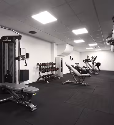 Et moderne treningsstudio med diverse treningsutstyr, inkludert en kabelmaskin, manualer på stativ, benker og kondisjonsmaskiner som elliptiske og stasjonære sykler. Rommet har sort gulv, hvite vegger, og synlig kanalføring i tak.