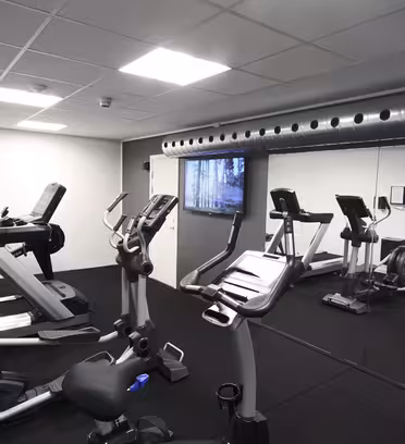 Et moderne innendørs treningsstudio med treningsutstyr, inkludert stasjonære sykler, tredemøller og en elliptisk trener. Rommet har speilvegg, mørkt gulv og ventilasjonsrør i metall langs taket.