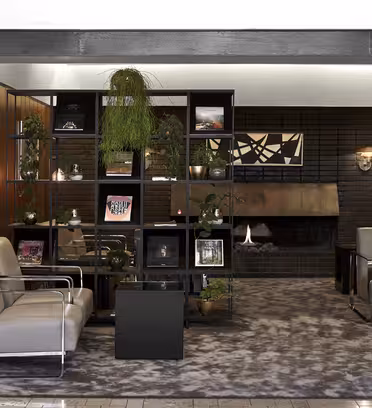 En moderne salong har elegante grå sitteplasser og en geometrisk hylle med planter og kunstverk. En peis er satt inn i en mørk murvegg, mens et teksturert teppe dekker gulvet. Varme tretoner fremhever den moderne innredningen.